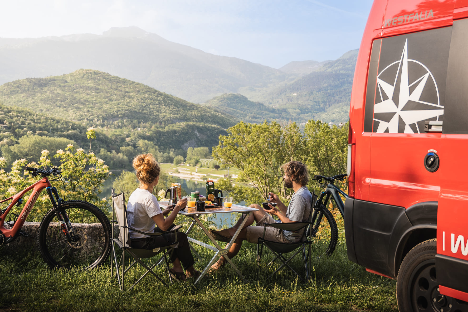 Zwei Personen sitzen an einem Tisch im Freien und genießen eine Mahlzeit neben einem roten Wohnmobil mit Bergen, Bäumen und einem See im Hintergrund. Zwei Fahrräder sind in der Nähe geparkt, und die Szene ist hell und friedlich.