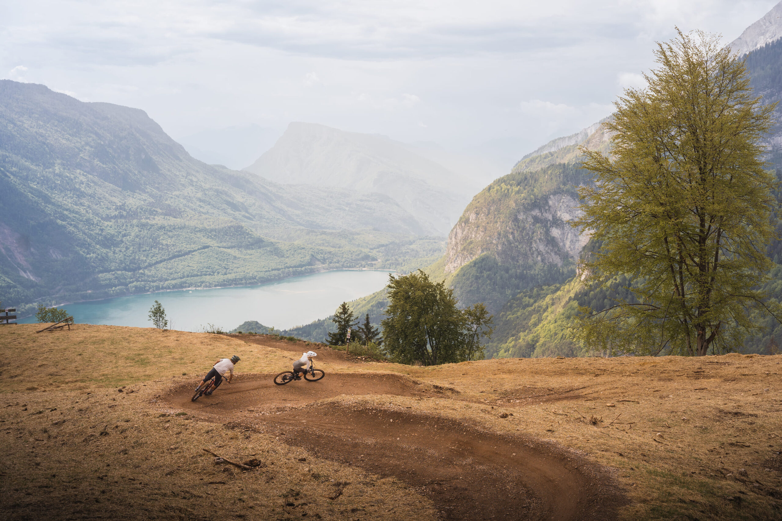 Zwei Mountainbiker fahren auf einem unbefestigten Weg an einem Hang mit Blick auf ein malerisches Tal, mit einem See und bewaldeten Bergen im Hintergrund unter einem bewölkten Himmel.
