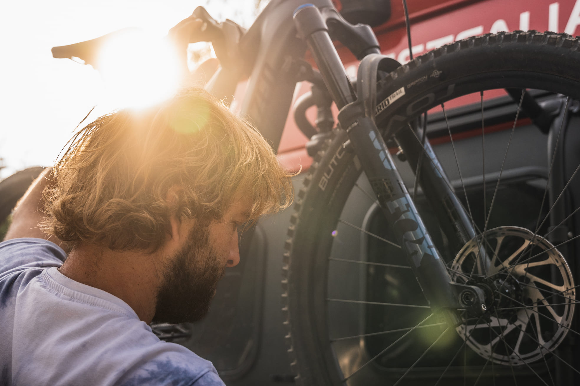 Ein Mann mit blondem Haar und Bart lädt bei Sonnenuntergang ein Mountainbike auf einen Gepäckträger, wobei das Sonnenlicht durch den Rahmen des Fahrrads scheint und im Hintergrund ein rotes Fahrzeug zu sehen ist.